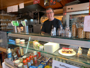 Der Kioskbetreiber hinter der Theke seines Lokals in der Mitte des Platzes am Hauptweg. Dort gibt es Brötchen, Croissant, Kuchen, Pizza, Currywurst mit hausgemachter Soße, Eis, Brötchenservice und Getränke aller Art